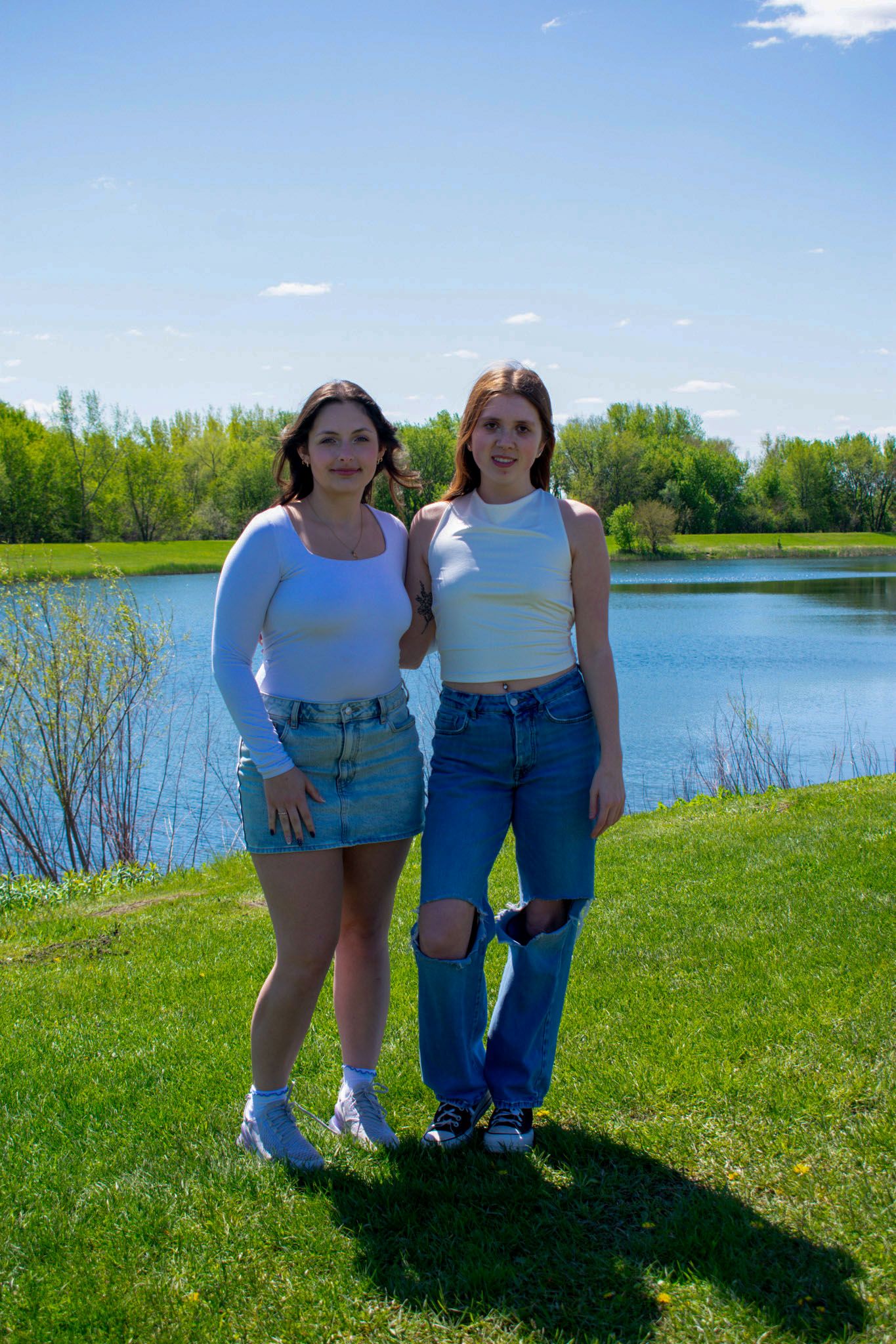 Two ladies with white tops and jean bottoms standing in front of a lake on sunny day