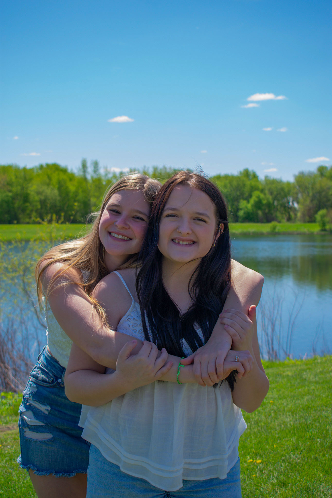 Two ladies with white tops and jean bottoms hugging each other in front of a lake on sunny day