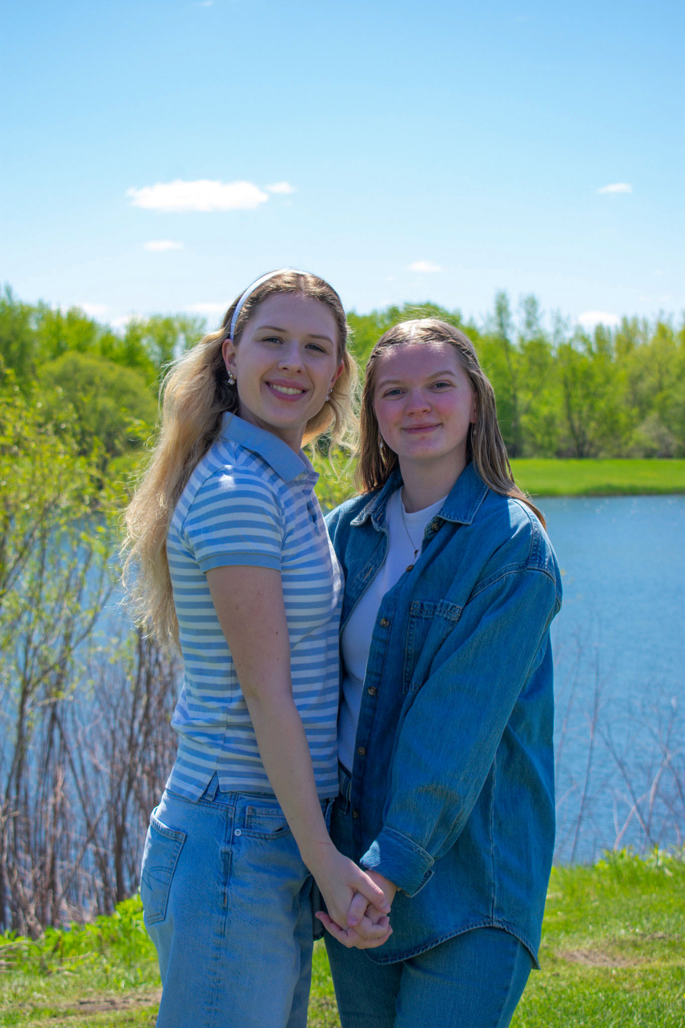 Two ladies with white tops and jean bottoms standing in front of a lake on sunny day