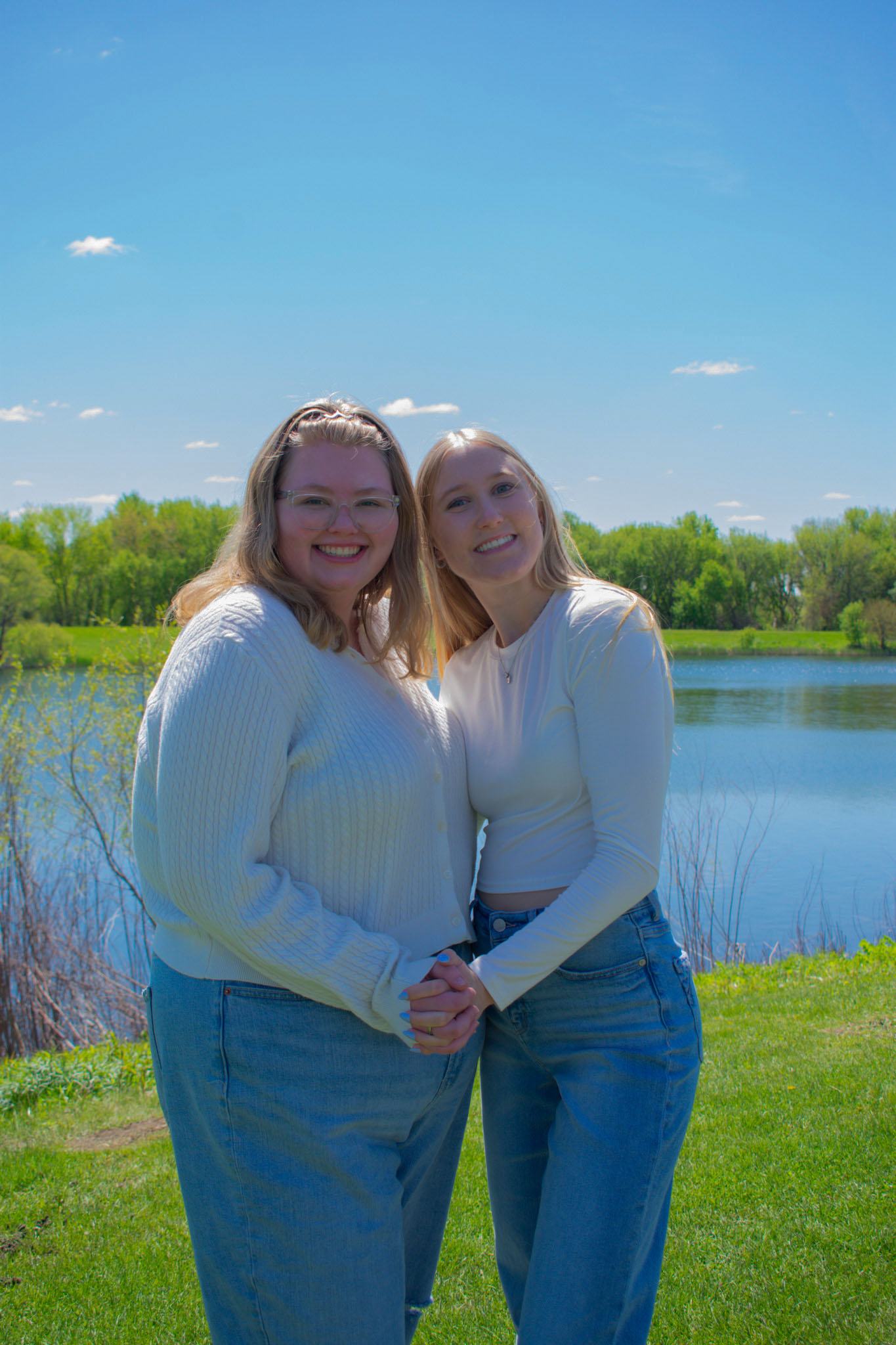Two ladies with white tops and jean bottoms standing in front of a lake on sunny day