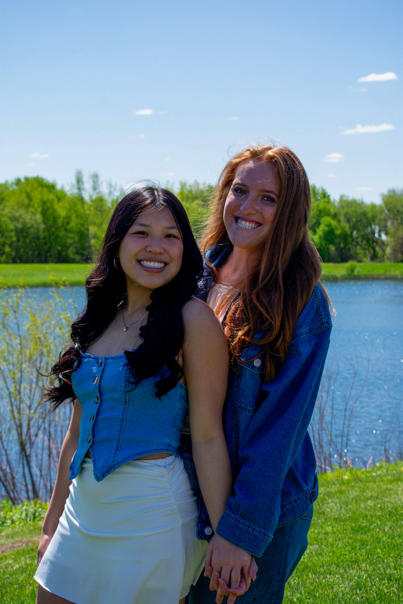 Two ladies with white tops and jean bottoms standing in front of a lake on sunny day