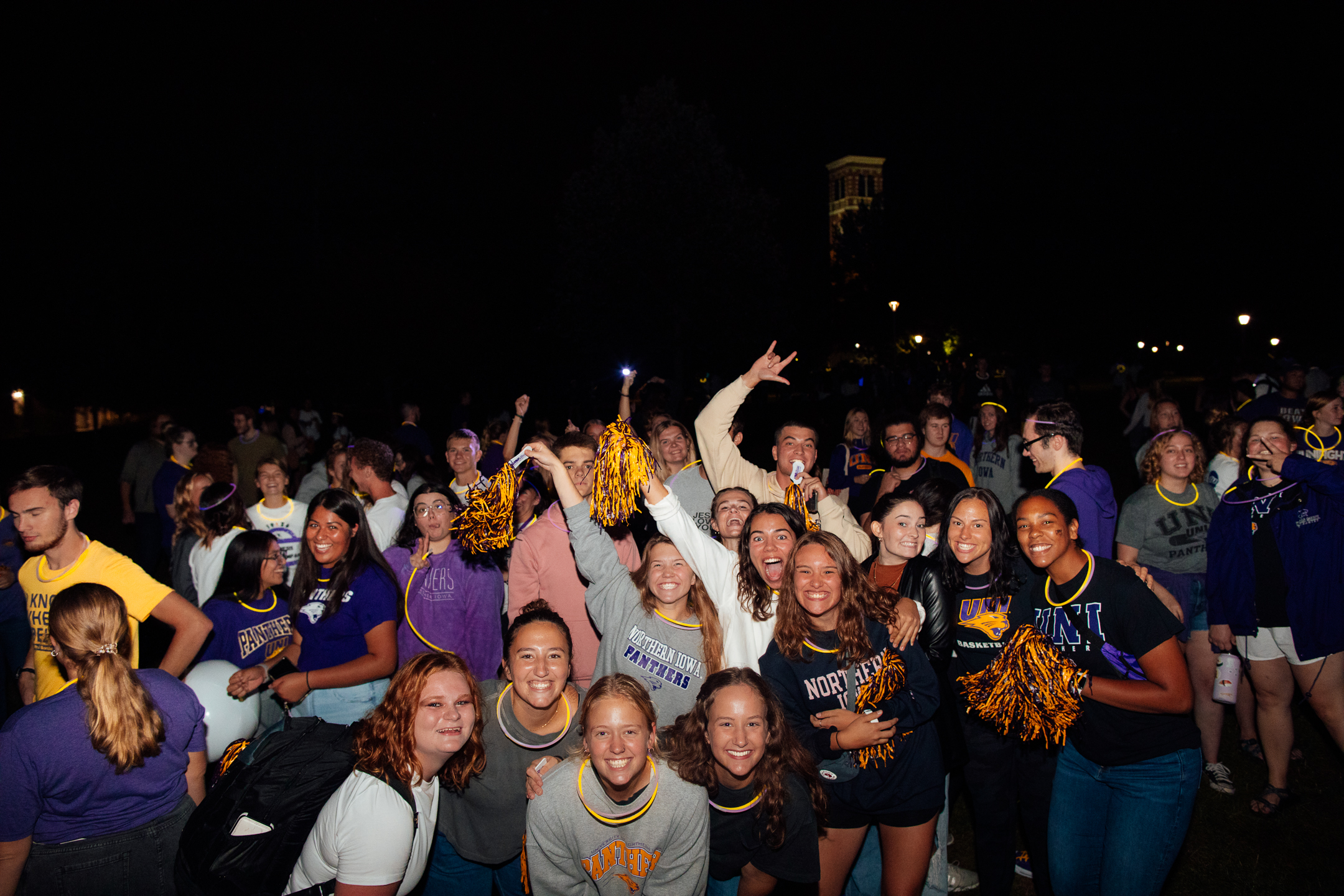 Exited and smiling students in the bottom half of image at night wearing UNI themed clothing and apparel. Dark black sky with the Campanile slightly lit in the background.