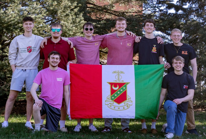 Group of gentlemen posing with their arms around each other with a Kappa Sigma Flag in front and pine trees behind them.