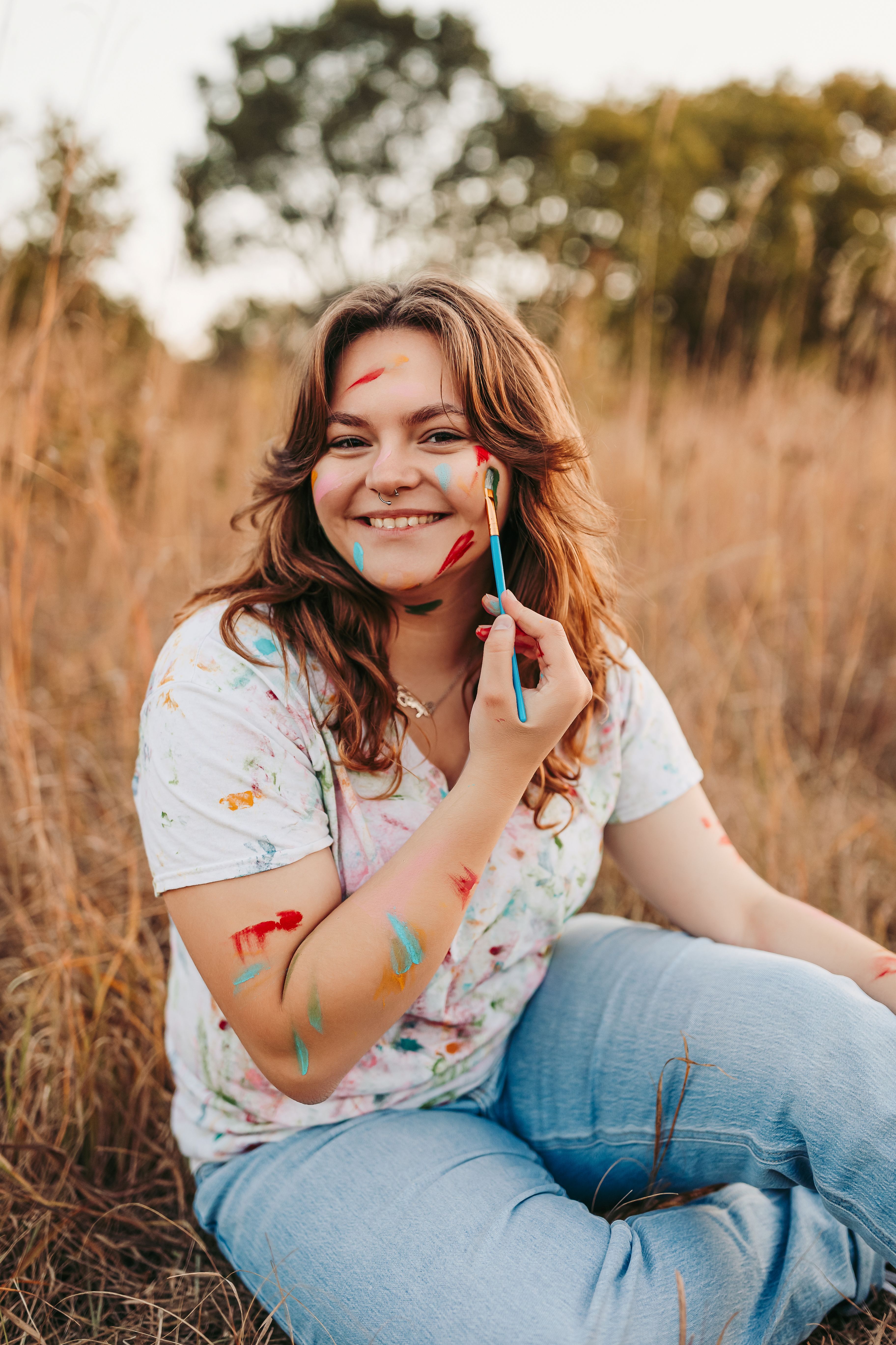 Young woman in a white shirt and jeans posing in a wheat field holding a paint brush with paint marks all over her